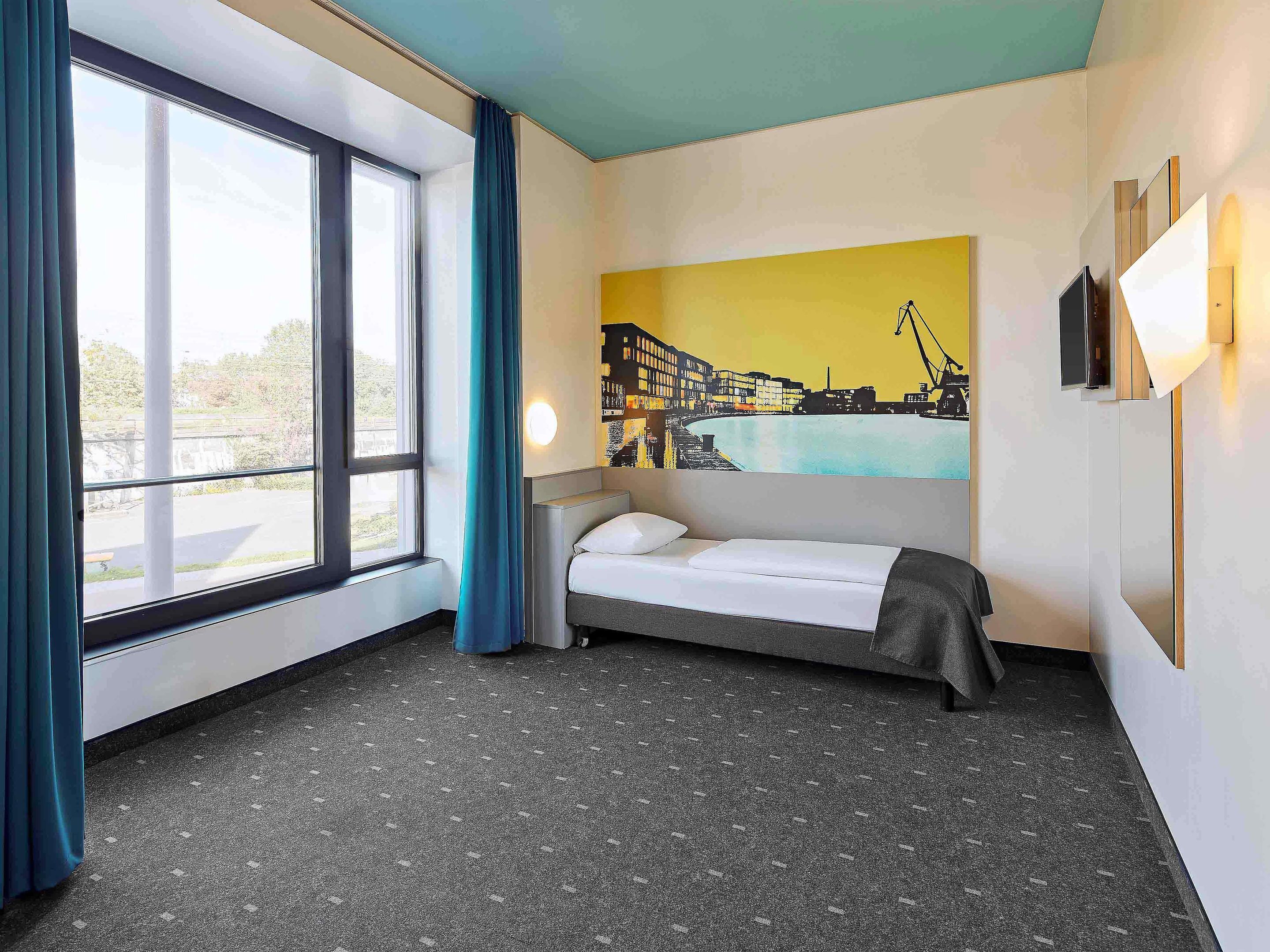 Habitación de hotel con cama, un ventanal con cortinas, un mural de paisaje urbano en la pared y un escritorio pequeño con lámpara.