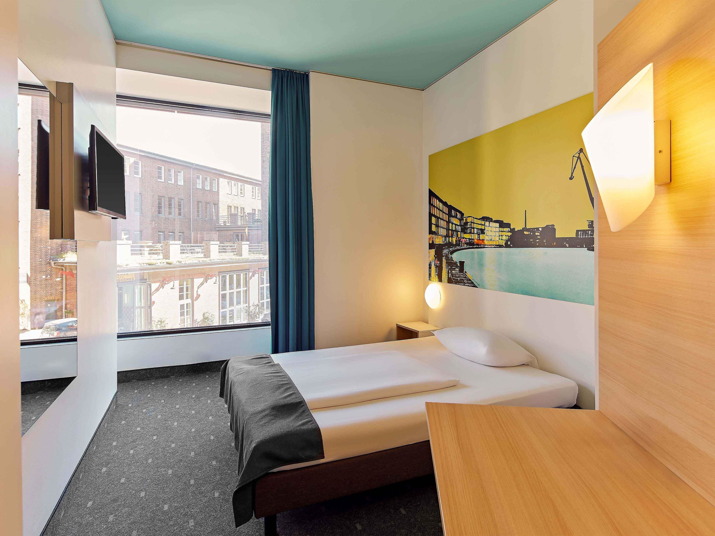 A hotel room with a bed, a desk, a window with a view of buildings, a painting on the wall, and a TV mounted on the wall.