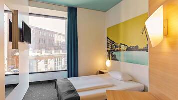 A hotel room with a bed, a desk, a window with a view of buildings, a painting on the wall, and a TV mounted on the wall.