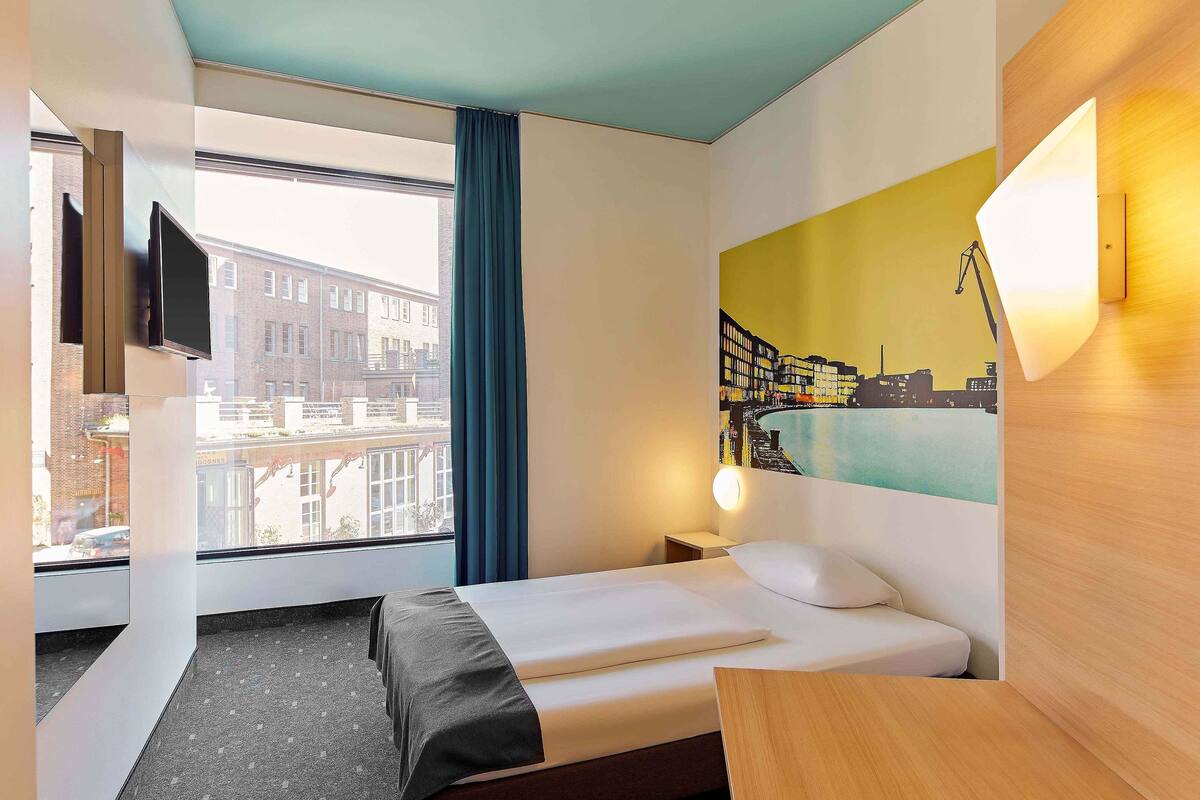 A hotel room with a bed, a desk, a window with a view of buildings, a painting on the wall, and a TV mounted on the wall.