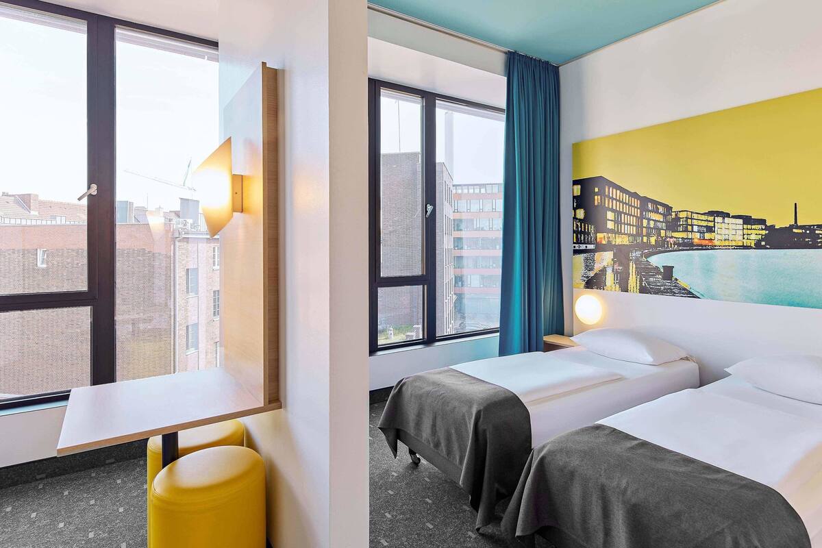 A hotel room with two beds, a desk, a chair, and a view of the cityscape.