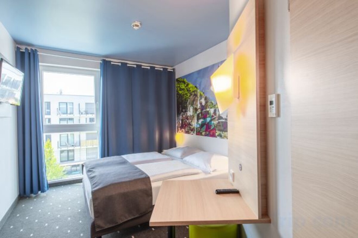 Habitación de hotel con cama, escritorio, silla, ventana con cortinas y una lámpara en la pared.