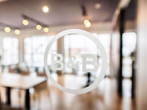 A circular logo with the letters "B&B" in a modern interior space with large windows and a blurred background. - B&B Hotel Neu-Ulm (Neu-Ulm)