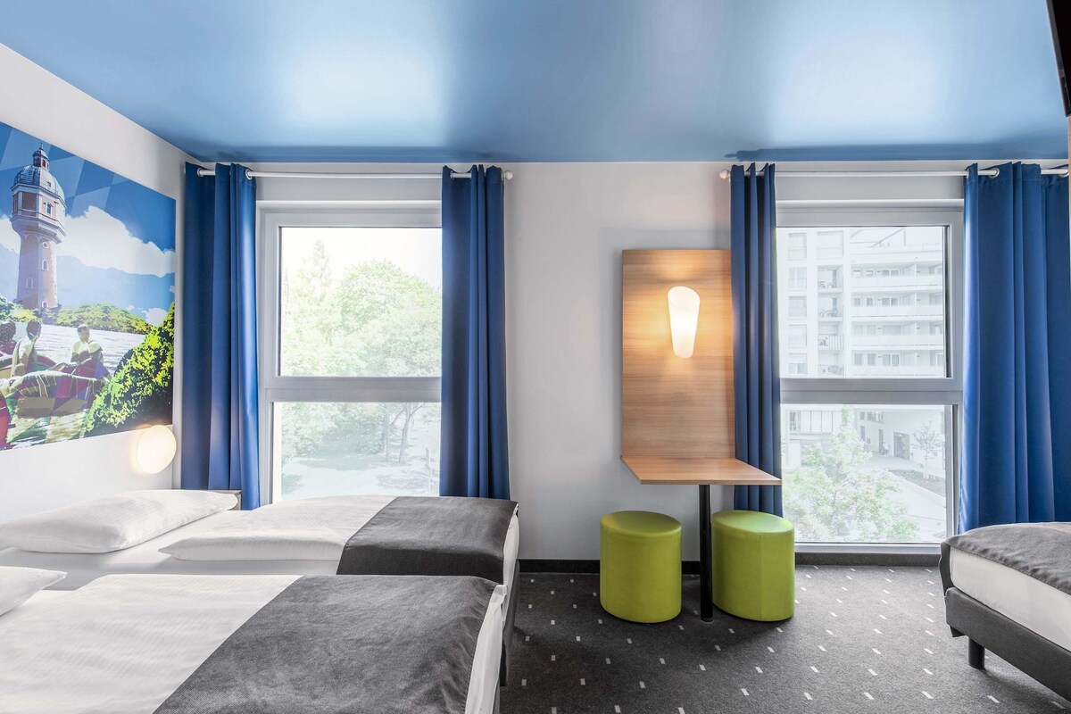 Une chambre d’hôtel avec deux lits, une lampe fixée au mur et une vue sur la ville grâce à de grandes fenêtres.