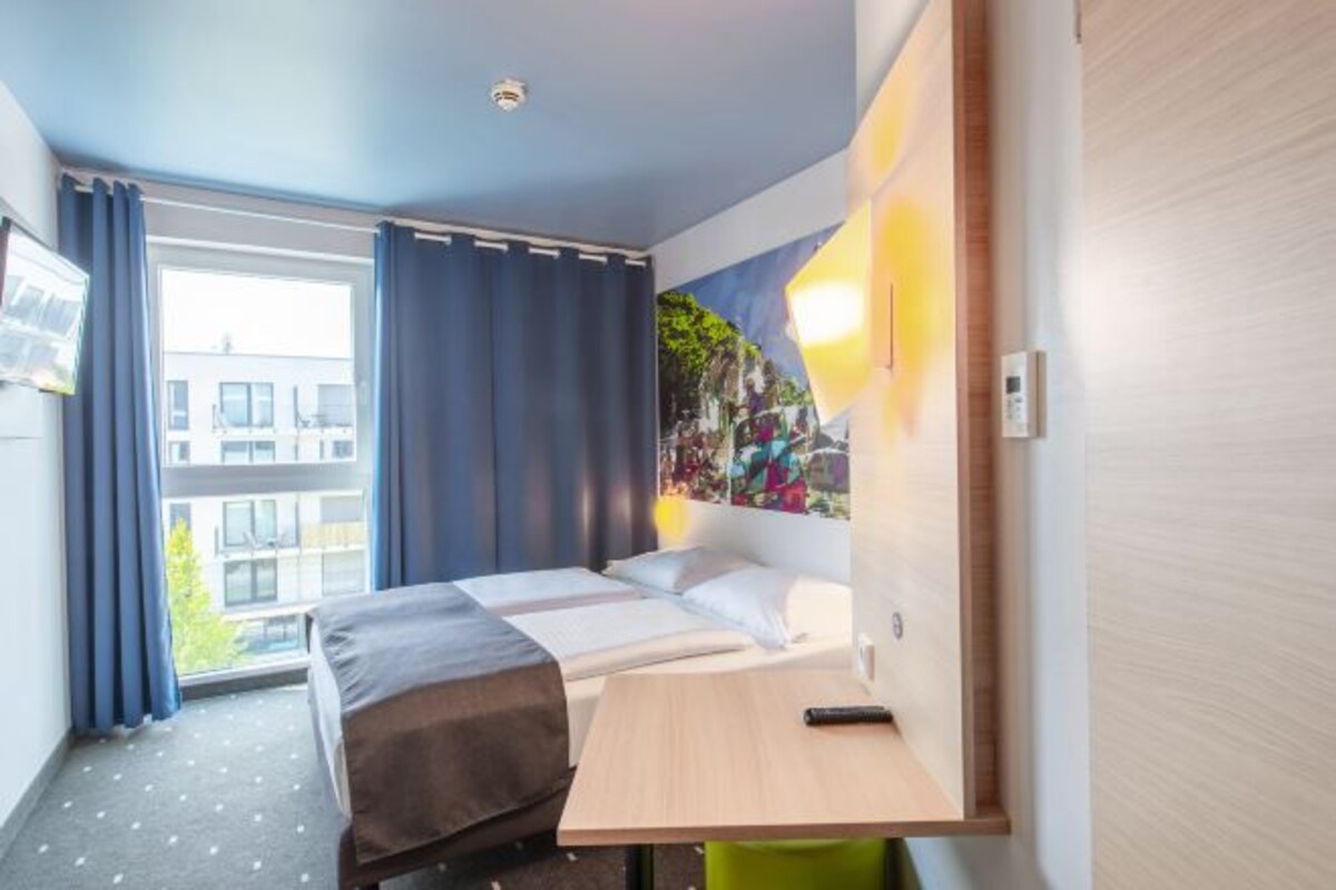 Ett hotellrum med en säng, ett skrivbord, en stol, ett fönster med gardiner och en vägglampa.