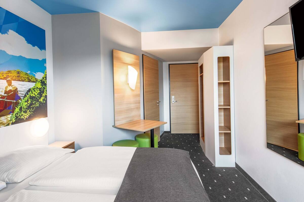 A modern hotel room with a bed, a desk, a wardrobe, and a large mirror.