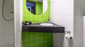 A bathroom with a green tiled wall, a round mirror, a sink, and a shower. - B&B Hotel Neu-Ulm (Neu-Ulm)
