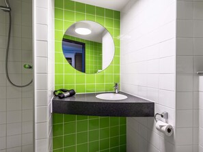 Une salle de bain avec un mur recouvert de carreaux verts, un miroir rond, un lavabo et une douche.