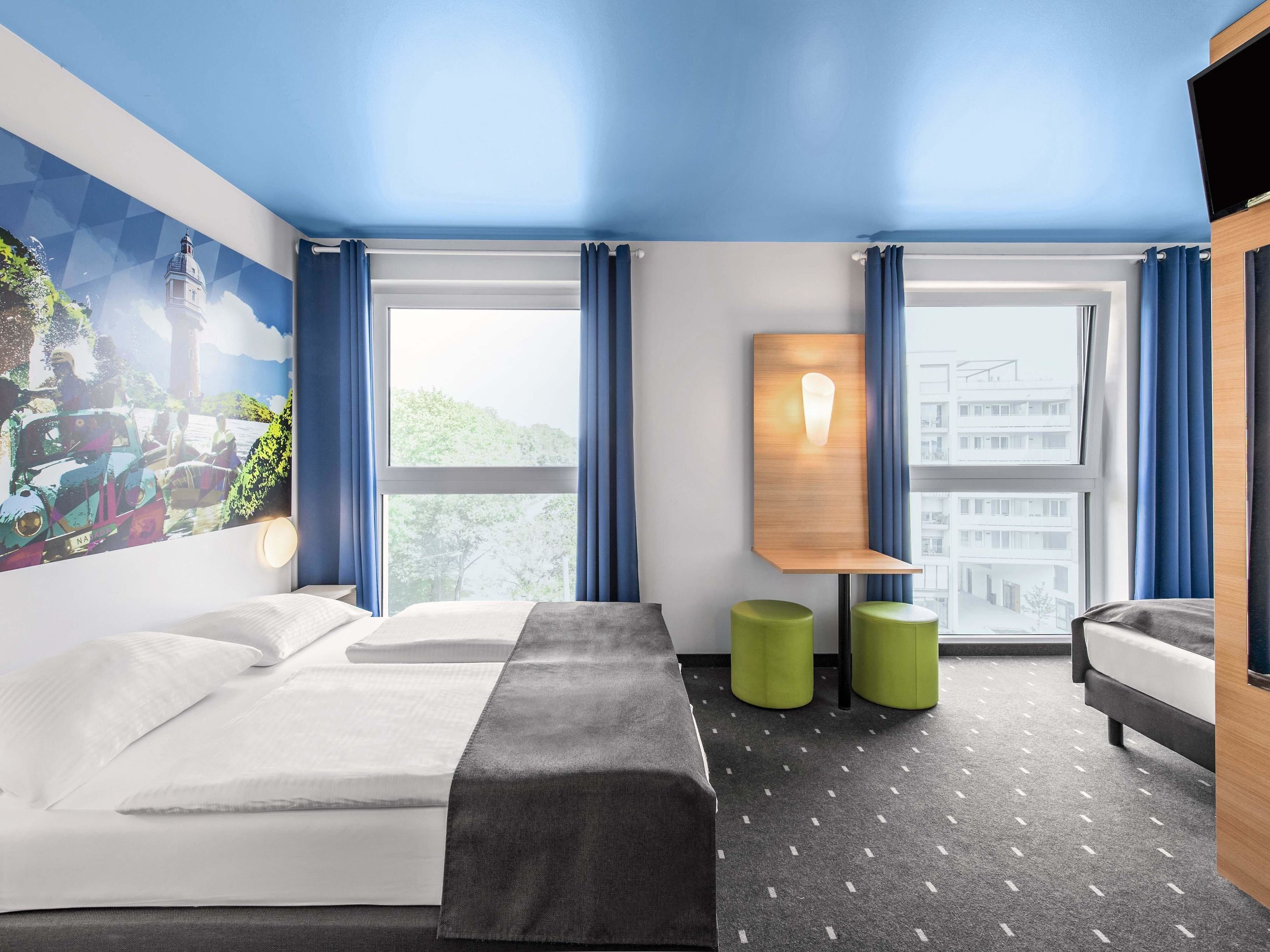 Ein Hotelzimmer mit zwei Betten, einem großen Fenster, einem Gemälde an der Wand und einem kleinen Tisch mit Lampe.