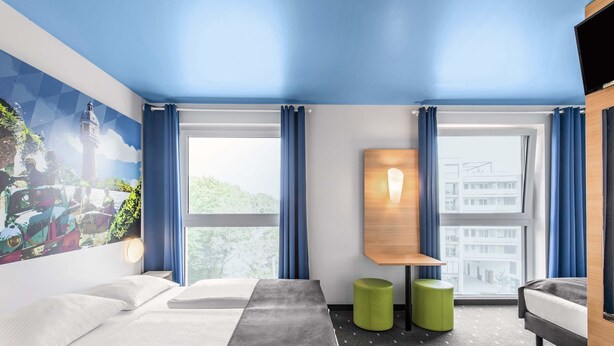 A hotel room with two beds, a large window, a painting on the wall, and a small table with a lamp. - B&B Hotel Neu-Ulm (Neu-Ulm)