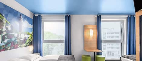 Ein Hotelzimmer mit zwei Betten, einem großen Fenster, einem Gemälde an der Wand und einem kleinen Tisch mit Lampe.