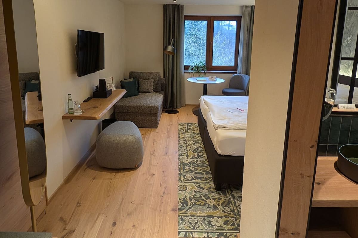 A compact hotel room with a bed, a small sofa, a desk with a chair, a TV mounted on the wall, and a window with a view of trees.