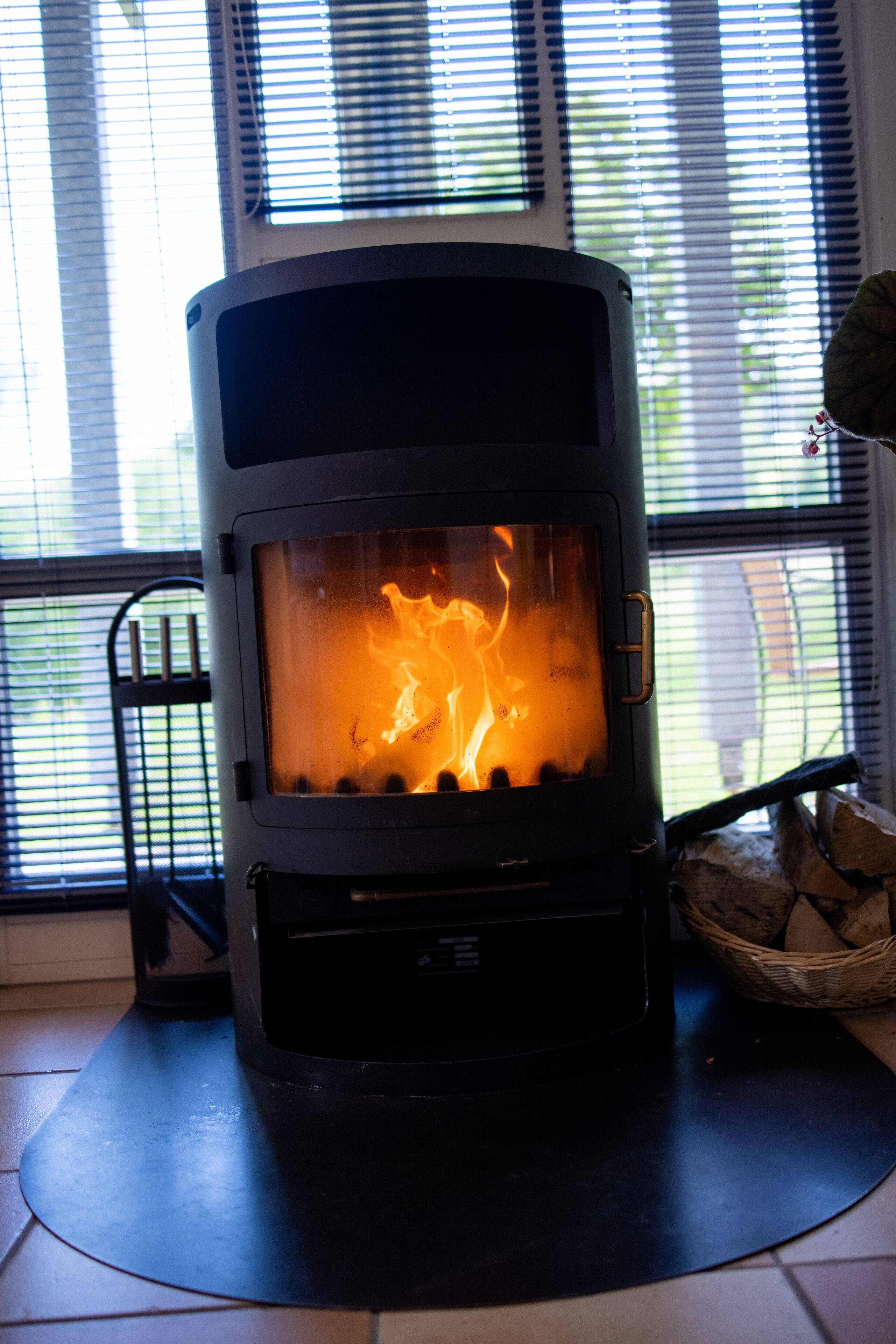 A wood-burning stove with a glass front, placed on a tiled floor near a window with blinds.