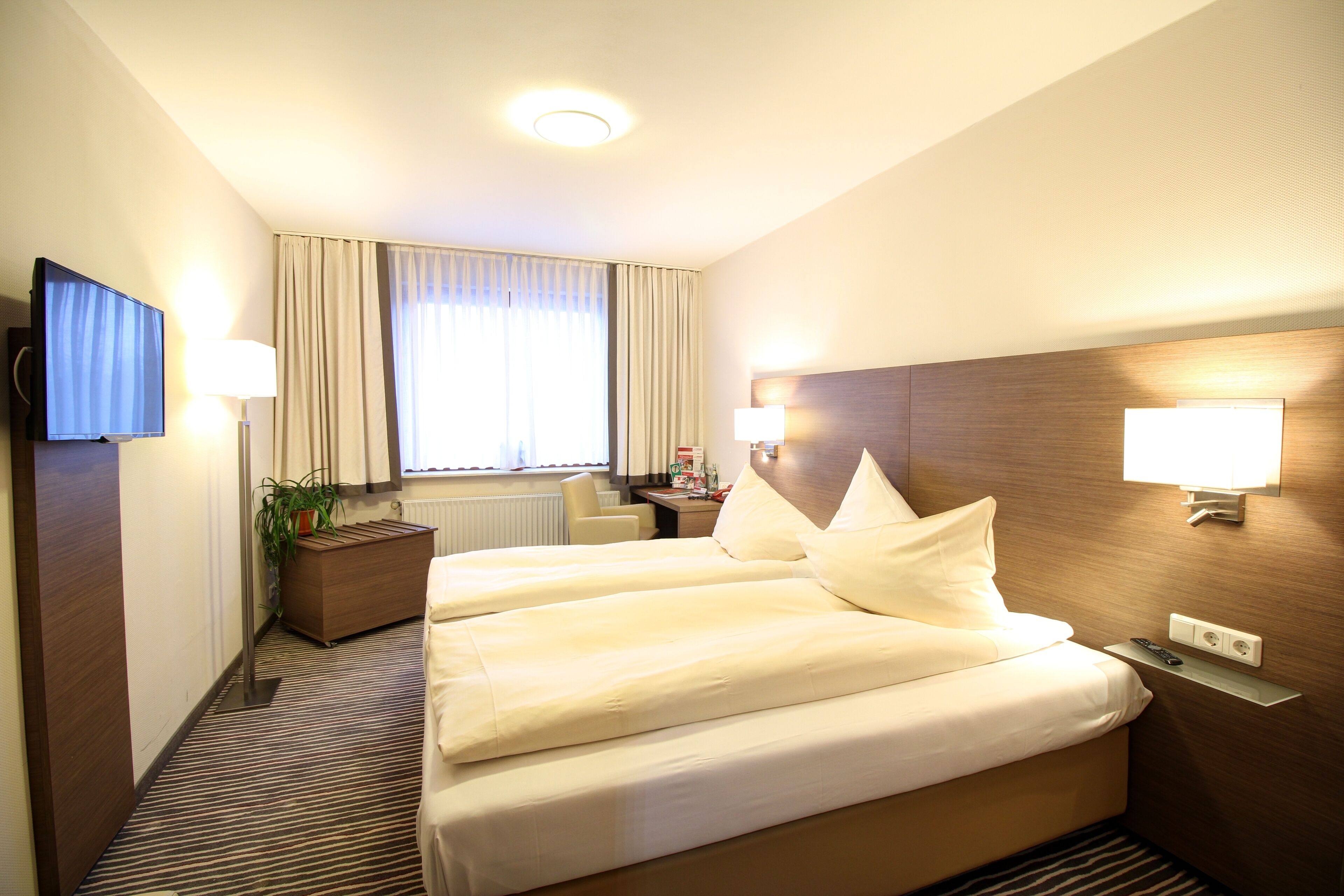 Habitación de hotel con una cama grande, lámparas de noche, un televisor de pantalla plana, un escritorio y una ventana con cortinas.