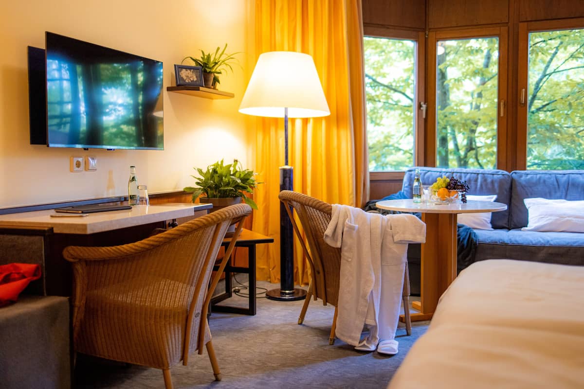 A hotel room with a flat-screen TV, a desk with a lamp, wicker chairs, a bed, and a view of trees through large windows.
