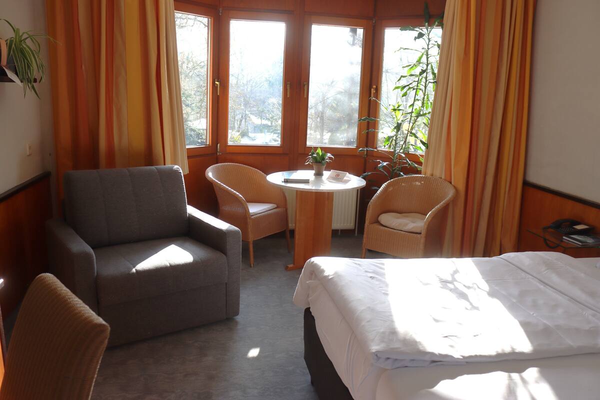 A hotel room with a bed, two armchairs, a small table, and a potted plant.