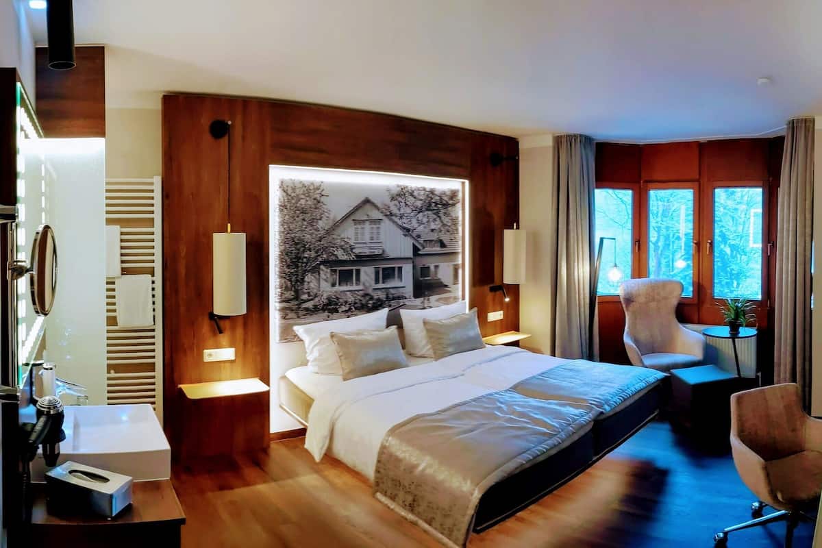 A hotel room with a large bed, a desk, a chair, a small table, a mirror, and a window with curtains.