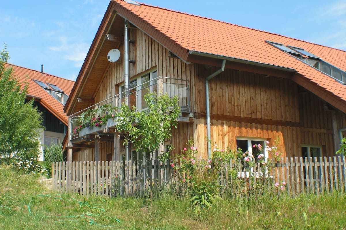 A wooden house with a red-tiled roof, a balcony with potted plants, and a wooden fence.
