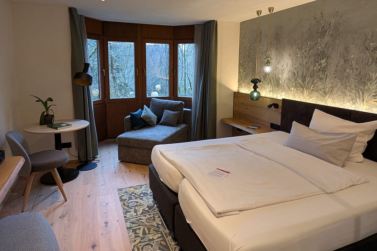 A hotel room with a large bed, a small sofa, a desk, and a view of trees through the windows.