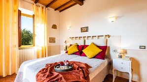 A neatly made bed with a red and yellow pillow set, a bedside table with a lamp, and a window with curtains. - Il Poggiarellino 14, Emma Villas (Volterra)