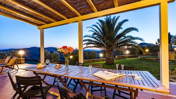 An outdoor patio with a wooden table and chairs, a vase of flowers, and a view of the hills. - Il Poggiarellino 14, Emma Villas (Volterra)