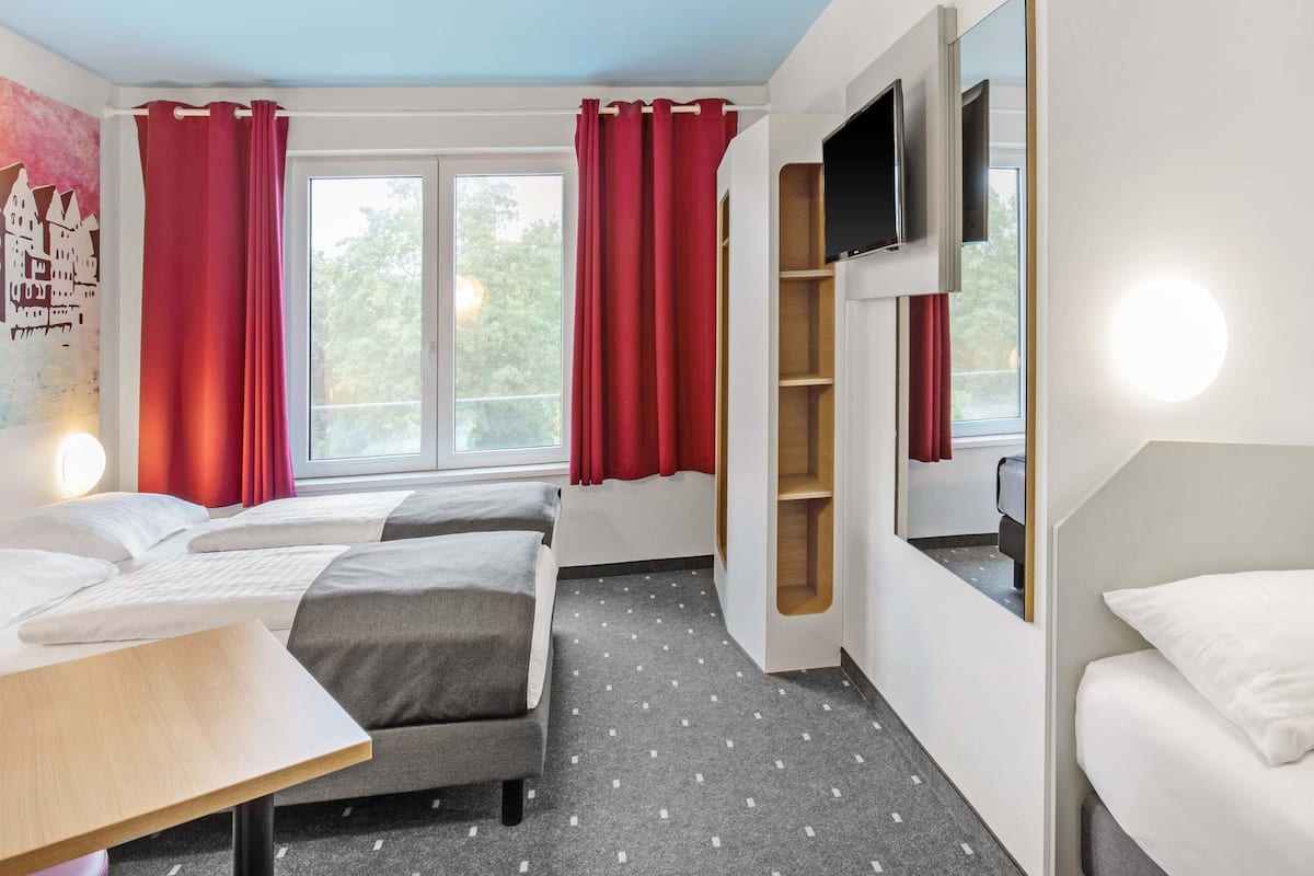 Habitación de hotel con dos camas, televisor, escritorio y ventana con cortinas rojas.
