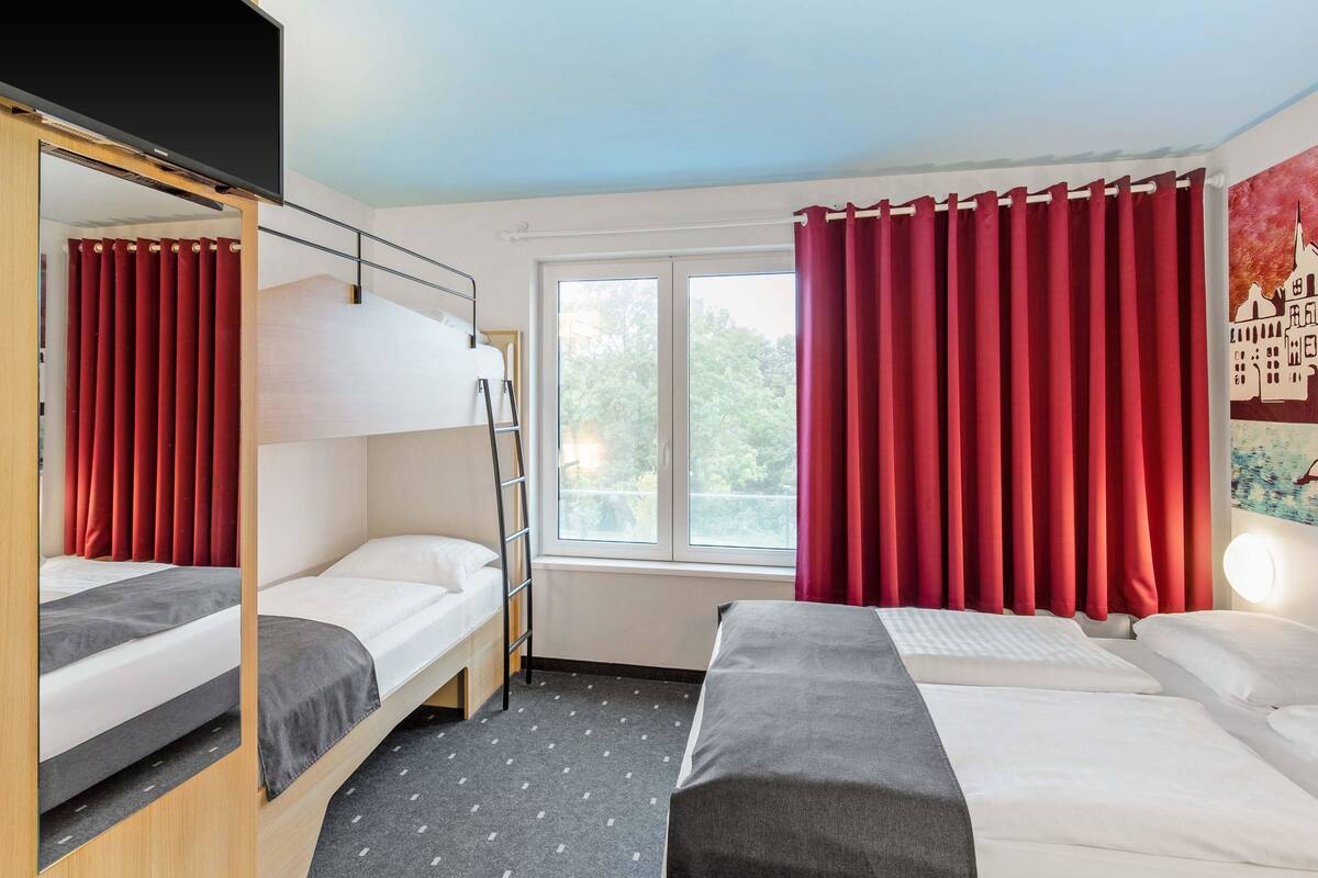 Una habitación con literas, una ventana con cortinas rojas y una cama con cabecera blanca.