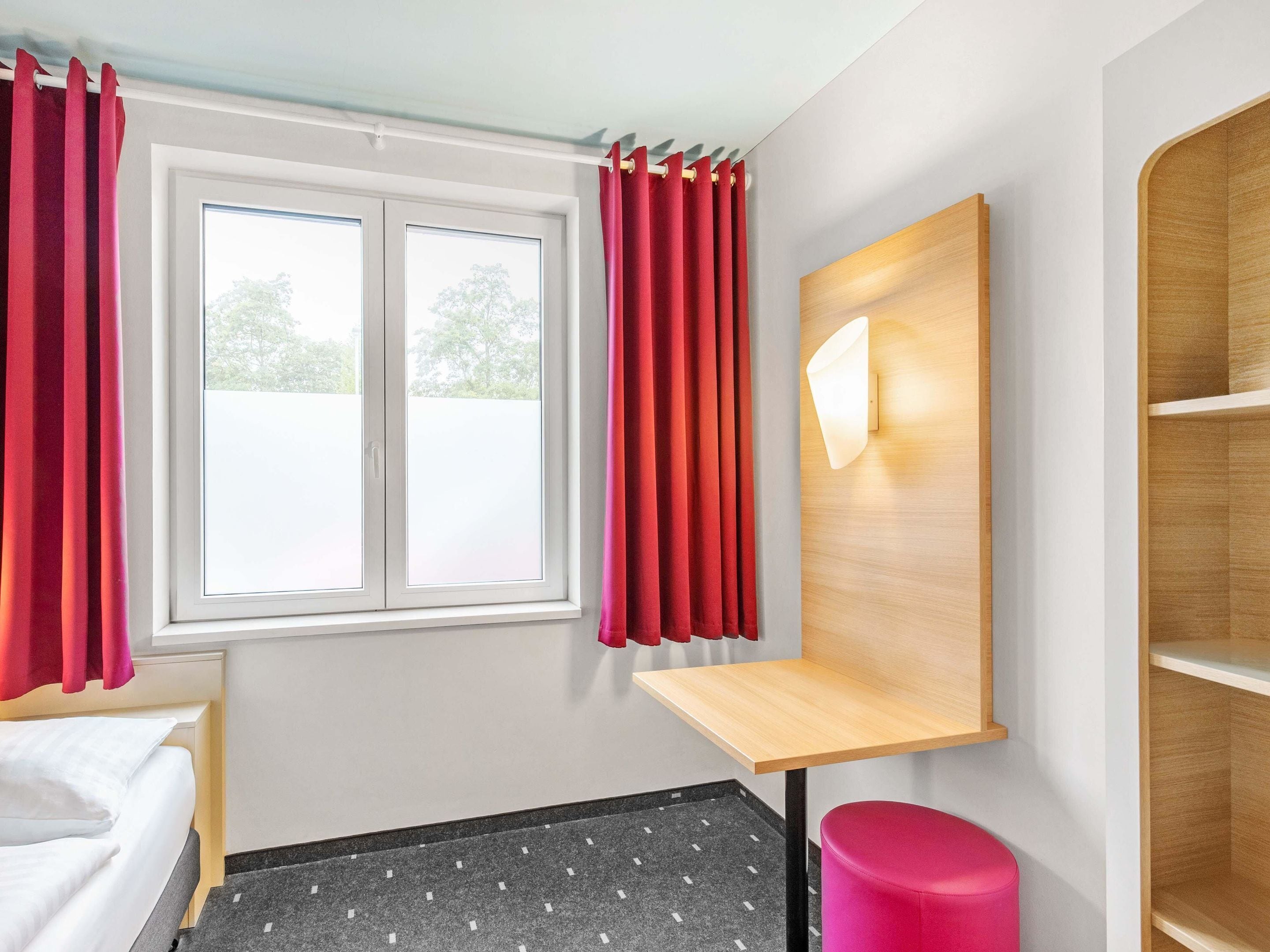 A hotel room with a bed, a desk, a chair, a window with curtains, and a wall-mounted shelf.