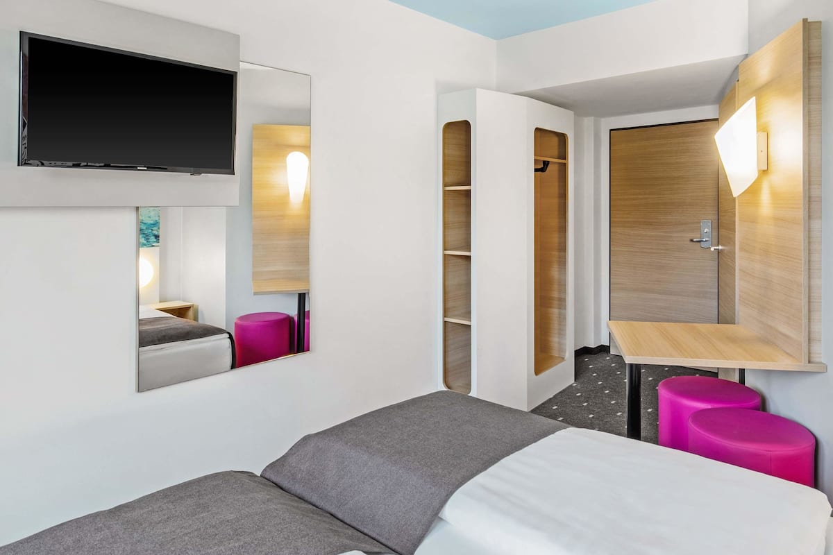 Una habitación de hotel moderna con una cama, un televisor montado en la pared, un armario de madera y un pequeño escritorio con una silla.