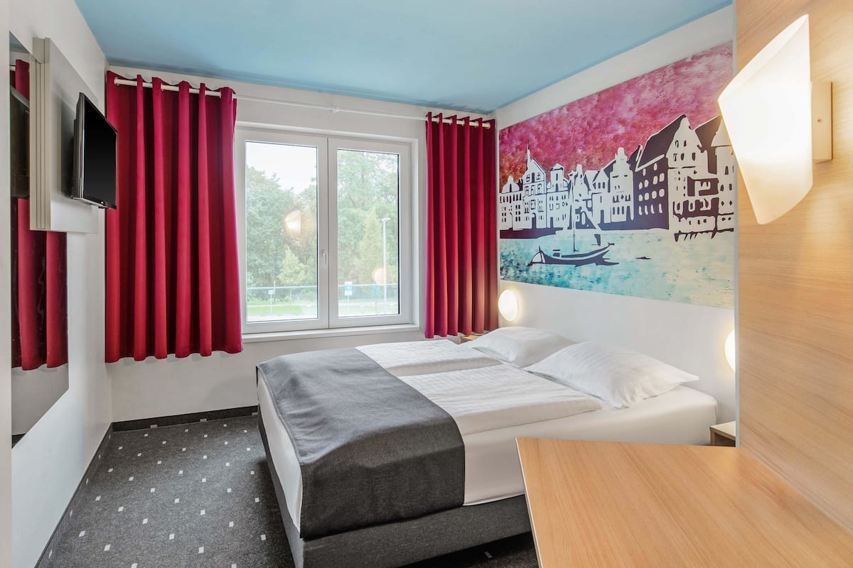 Habitación de hotel con cama, televisión, ventana con cortinas rojas y un cuadro de un paisaje urbano en la pared.