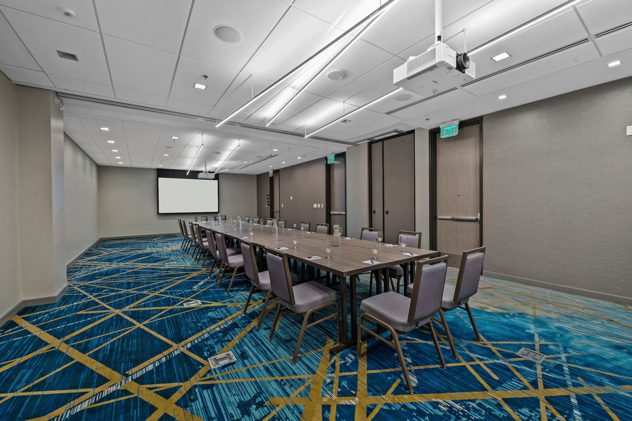 A conference room with a long table, chairs, a projector screen, and a carpeted floor.