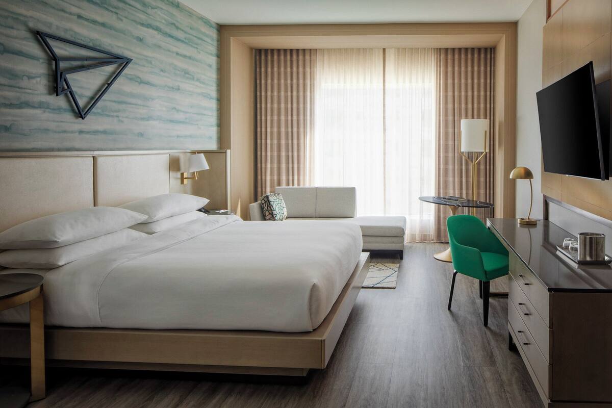 Habitació d'hotel amb un llit gran, una taula amb cadira, una televisió i una finestra amb cortines.