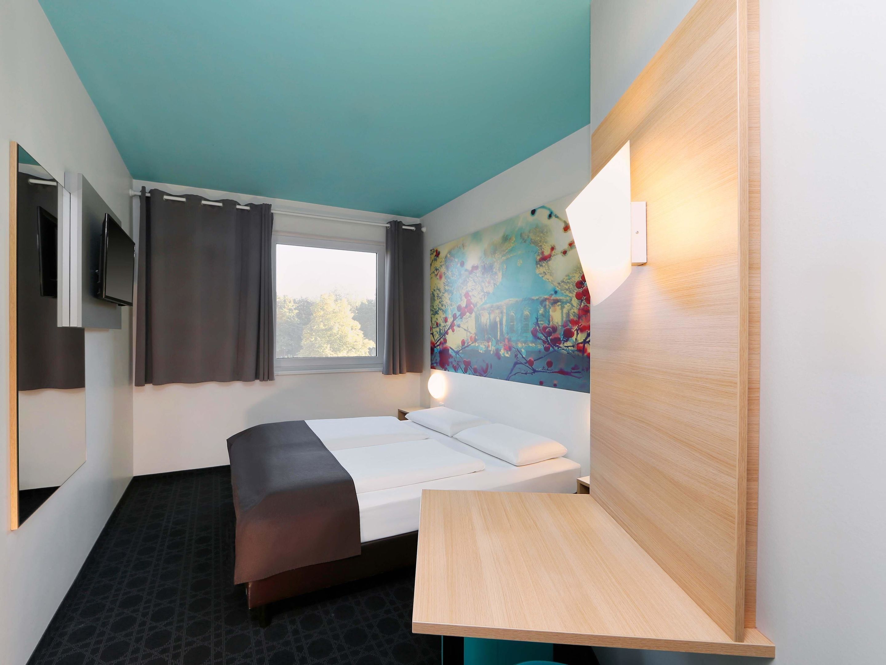 A modern hotel room with a bed, a desk, a TV, and a window with curtains.