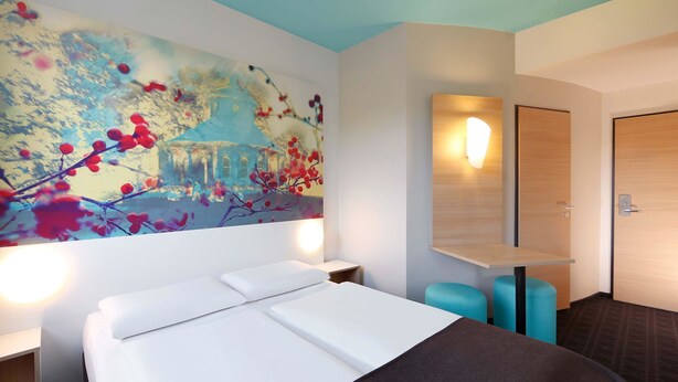 A hotel room with a large bed, a wall-mounted TV, a bedside table, a nightstand, and a door with a light switch. - B&B Hotel Potsdam (Potsdam)