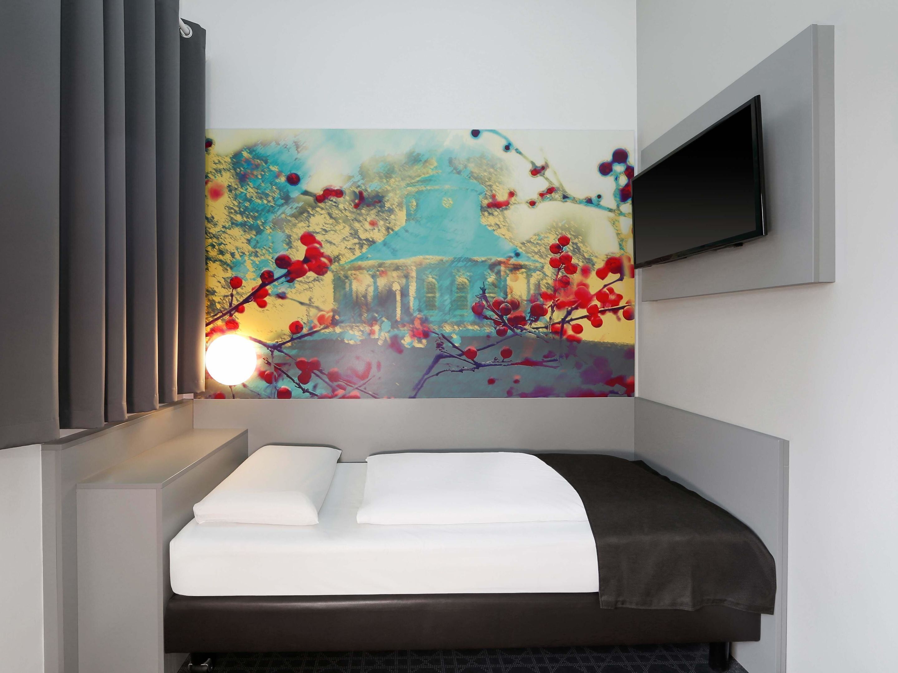 A modern hotel room with a bed, a flat-screen TV mounted on the wall, and a large abstract painting above the bed.