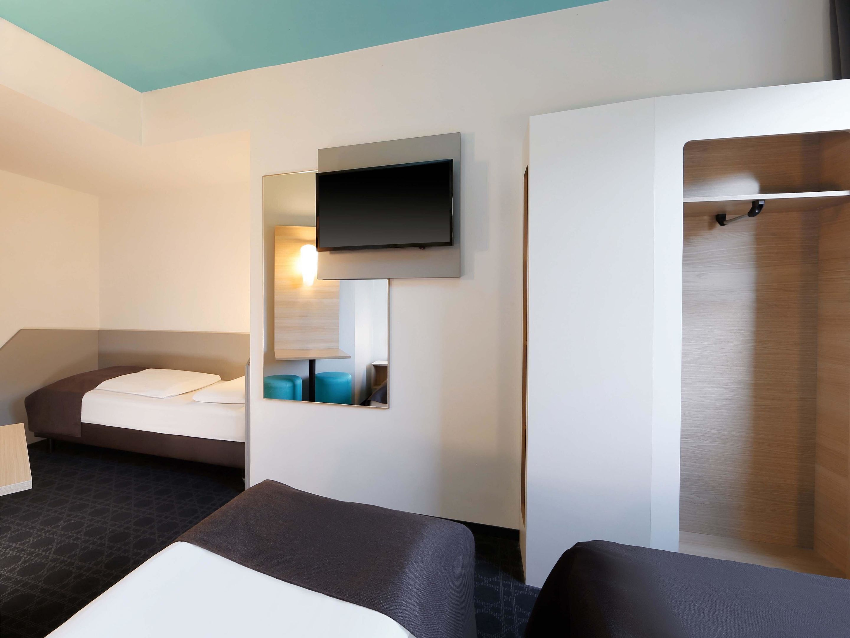 A modern hotel room with a bed, a sofa, a TV mounted on the wall, and a mirror reflecting the room.