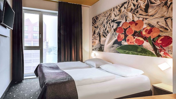 A hotel room with a bed, a TV mounted on the wall, a large window with curtains, and a wall with a floral painting. - B&B Hotel Bremen-Überseestadt (Bremen)