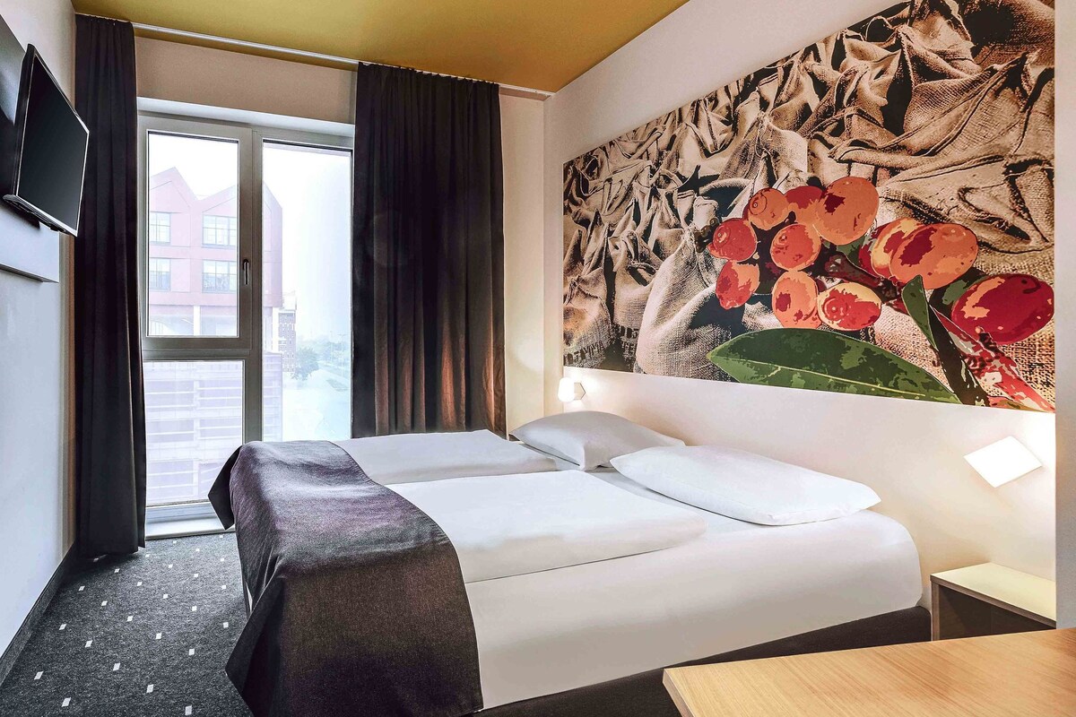 A hotel room with a bed, a TV mounted on the wall, a large window with curtains, and a wall with a floral painting.