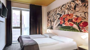 A hotel room with a bed, a TV mounted on the wall, a large window with curtains, and a wall with a floral painting.