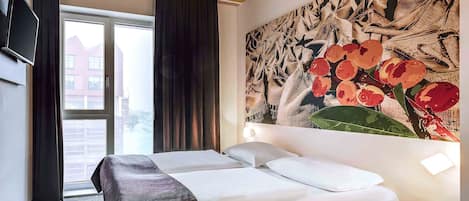 Una habitación de hotel con una cama, un televisor montado en la pared, un ventanal con cortinas y un cuadro floral.
