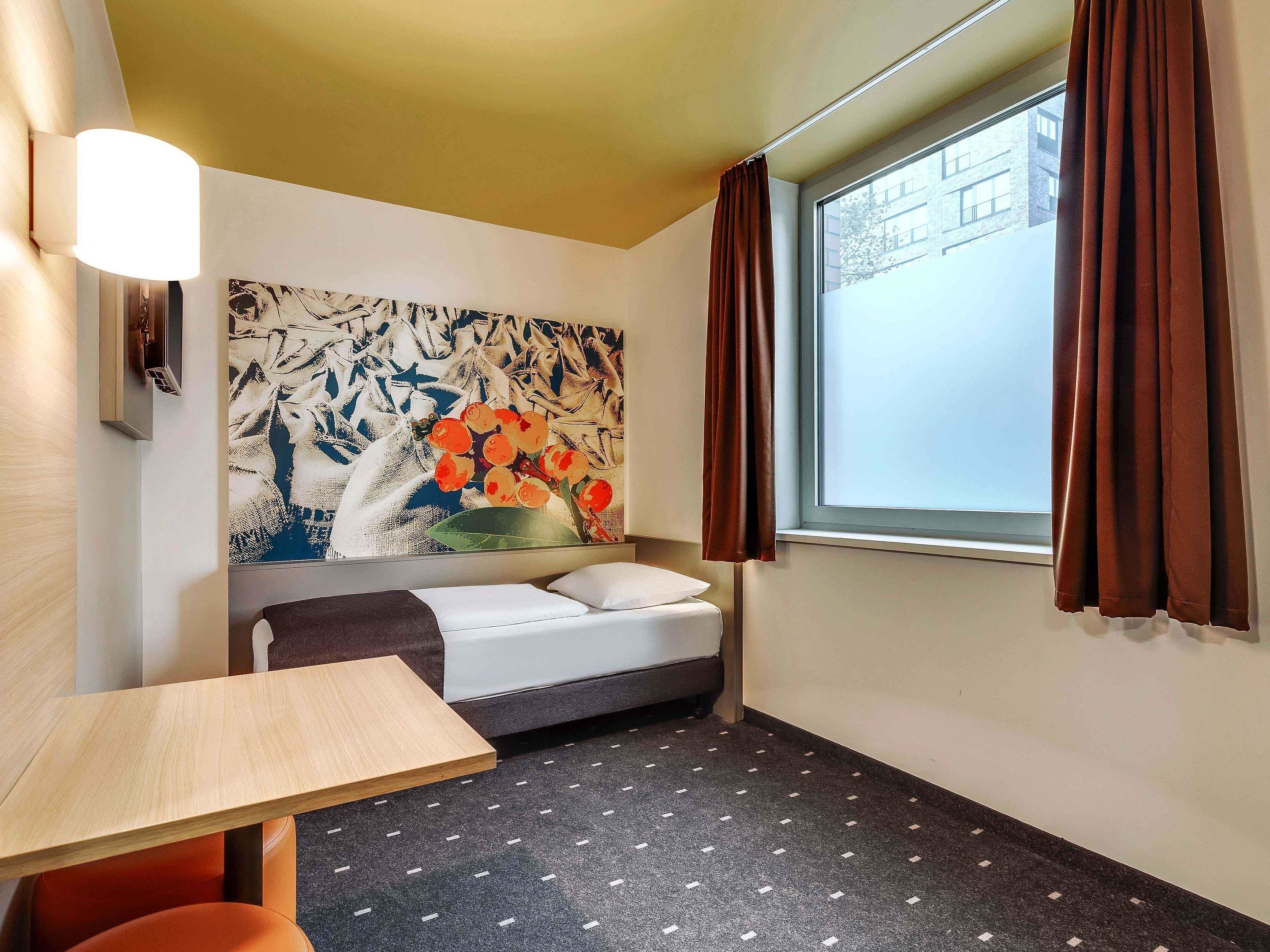 Habitación de hotel con cama, una mesita, luz empotrada en la pared, ventana con cortinas y un cuadro abstracto de gran tamaño.