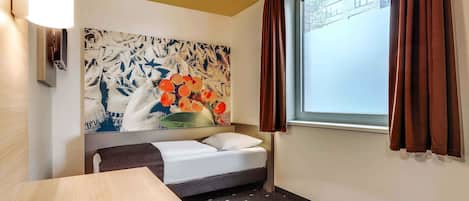 Une chambre d’hôtel avec un lit, une petite table, une lampe fixée au mur, une fenêtre avec des rideaux et un grand tableau abstrait.