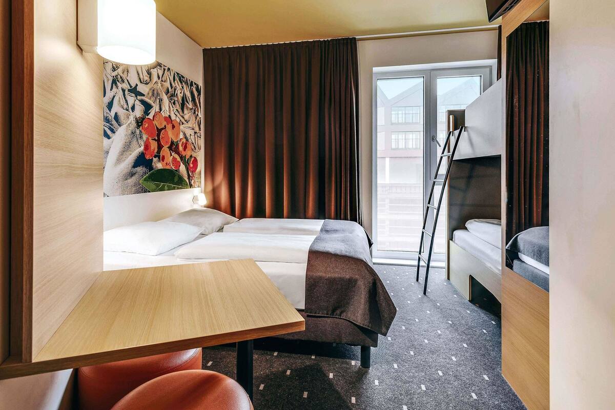 A hotel room with a bed, a desk, a chair, a painting, and a view of the outside.