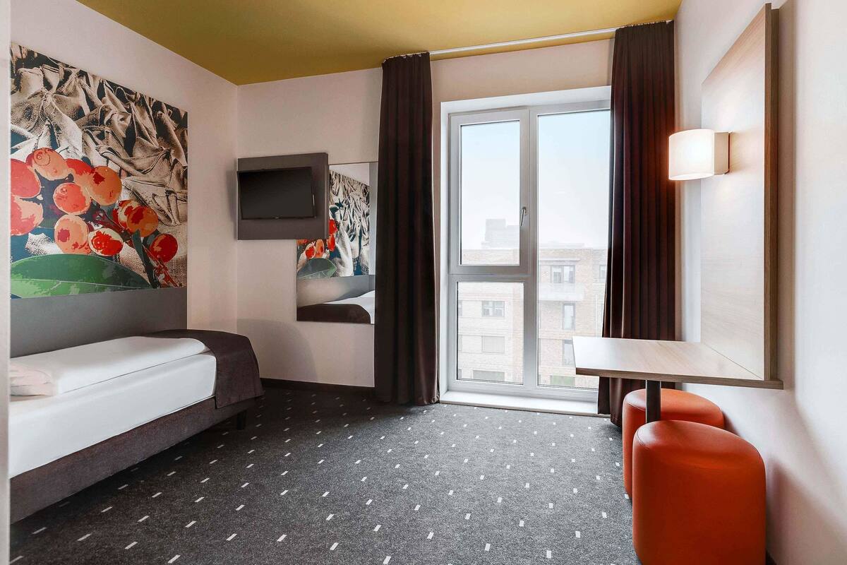 A hotel room with a bed, a TV mounted on the wall, a window with curtains, and a small table with a chair.