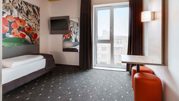 A hotel room with a bed, a TV mounted on the wall, a window with curtains, and a small table with a chair. - B&B Hotel Bremen-Überseestadt (Bremen)