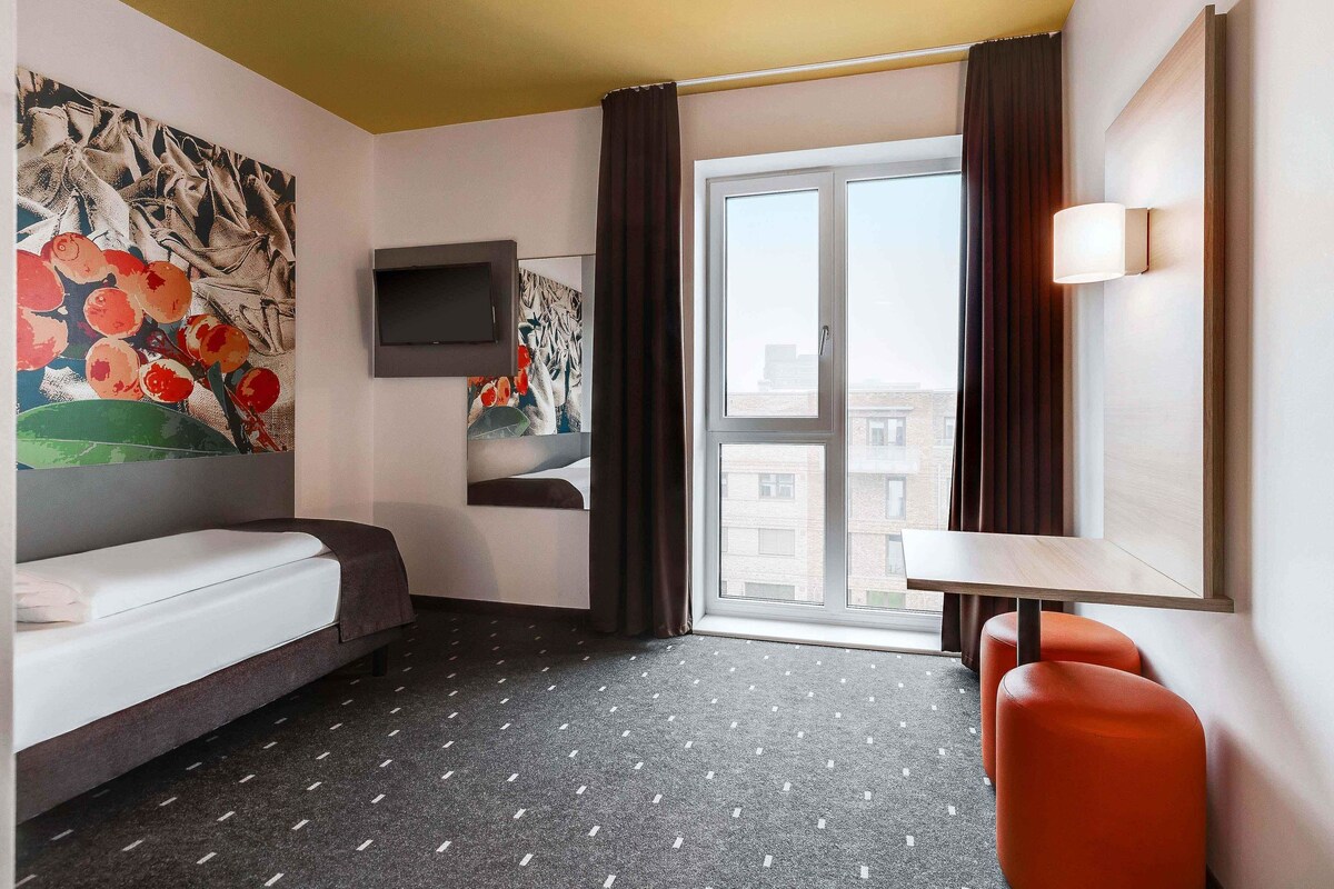 A hotel room with a bed, a TV mounted on the wall, a window with curtains, and a small table with a chair.