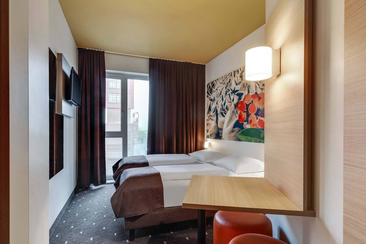 A hotel room with a bed, two bedside tables, a desk, a chair, a TV, and a window with curtains.