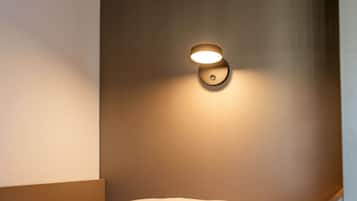 Una cama bien hecha con cabecera frente a una pared lisa, un aplique de pared y un objeto circular pequeño en la pared.