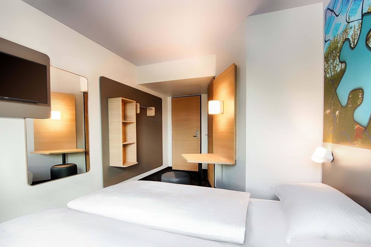 A modern hotel room with a large bed, a built-in desk, a mirror, and a wall-mounted light fixture.