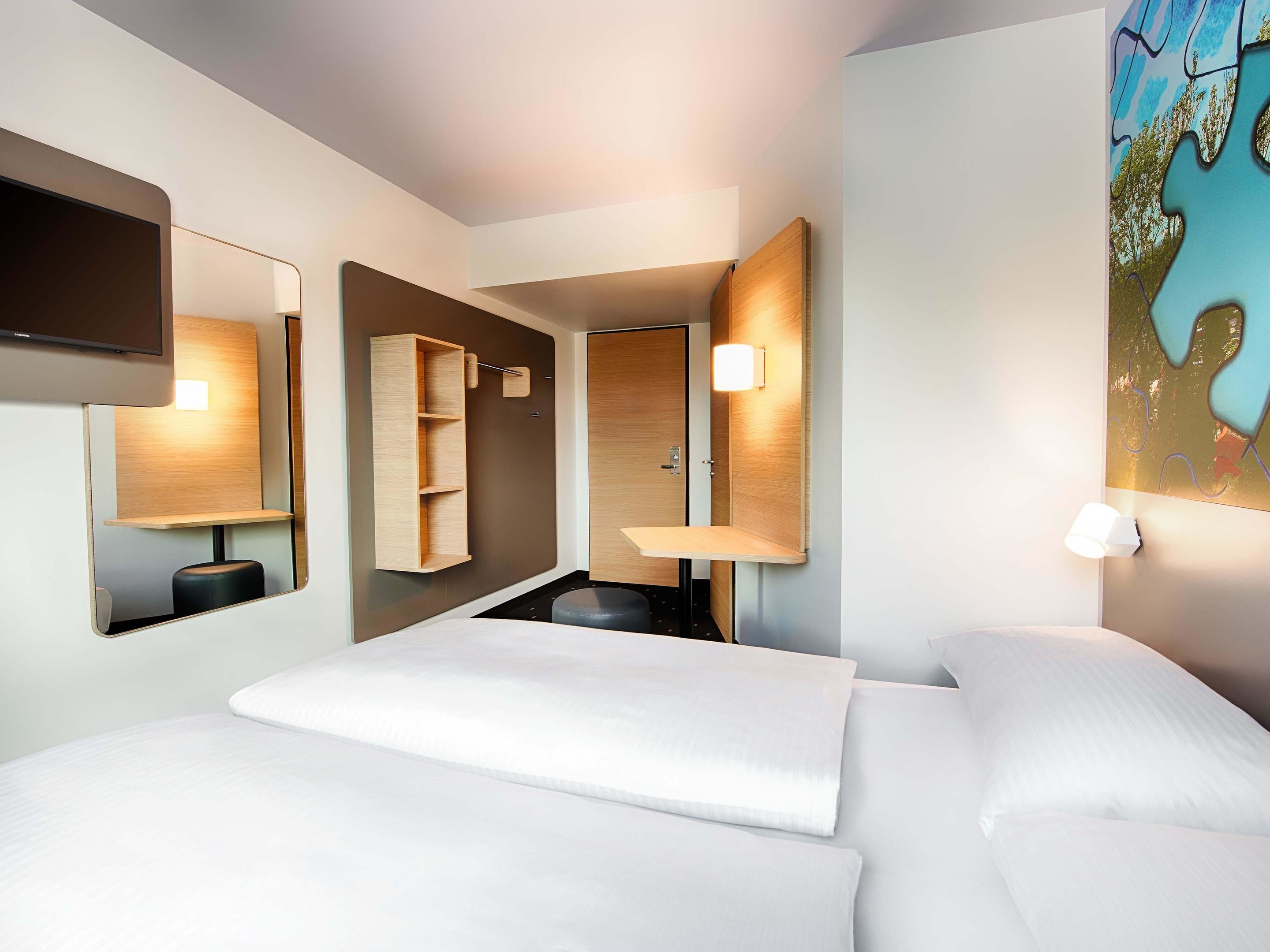 A modern hotel room with a large bed, a built-in desk, a mirror, and a wall-mounted light fixture.
